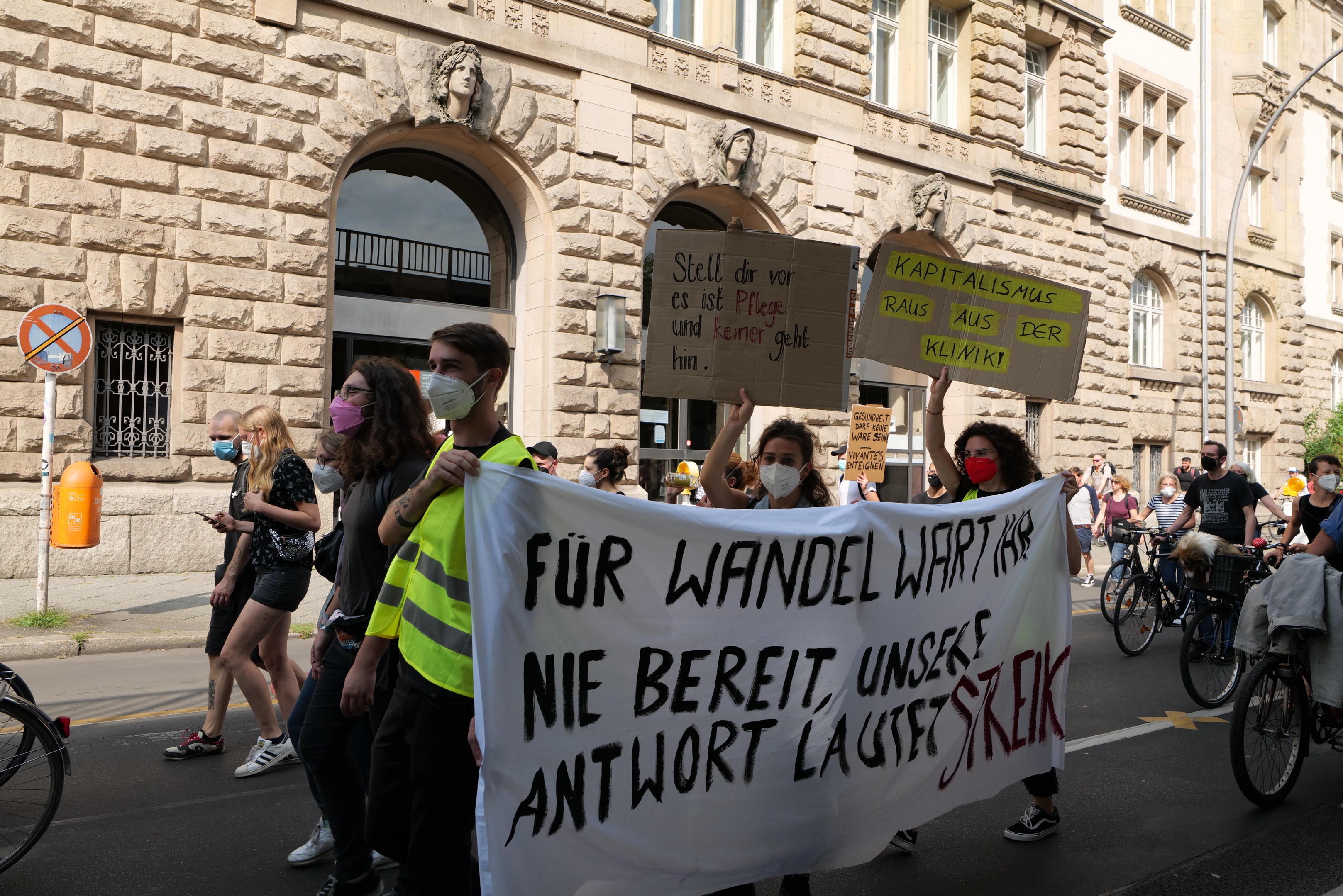 Eine Gruppe von Menschen, die bei einer Demonstration durch die Straße marschieren, einige halten Schilder und andere fahren Fahrräder, im Hintergrund ein Gebäude mit Fenstern, Bogengängen, Säulen und Skulpturen.