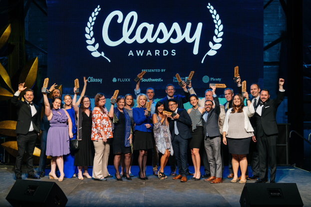 Eine Gruppe von Menschen auf einer Bühne, die lächeln und Auszeichnungen halten, mit Lautsprechern unten und einem Hintergrundbild mit der Aufschrift "Classy Awards 2019".