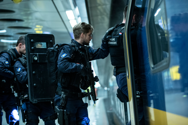Eine Gruppe von Polizisten steht neben einem Zug auf der rechten Seite des Bildes, wobei einer der Polizisten eine Waffe hält und an der Decke Lampen befestigt sind.