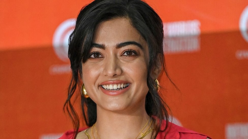 Eine Frau in einem roten Kleid mit einem eleganten Dutt und dezentem Make-up lächelt in die Kamera, trägt schwarze High Heels und hat ein hellgelbes Banner mit der Aufschrift 'Sonam Kapoors beste Schönheits Looks vom Cannes Film Festival' im Hintergrund.
