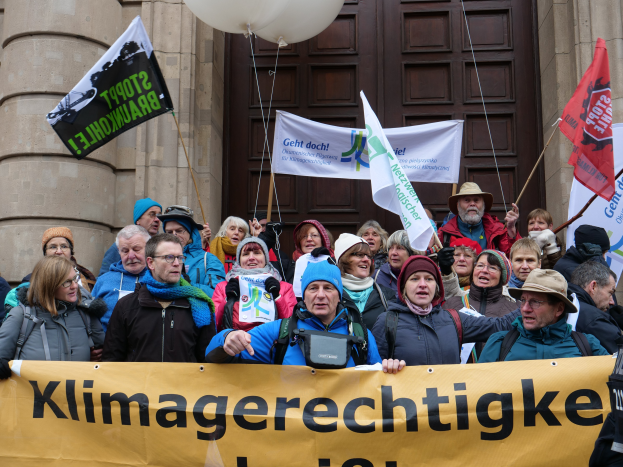 Eine Gruppe von Menschen, die vor einem Gebäude mit Säulen und einer Tür stehen und ein Transparent mit Text halten, sowie Fahnen und Luftballons, nehmen an einem Klimastreik in Deutschland teil.