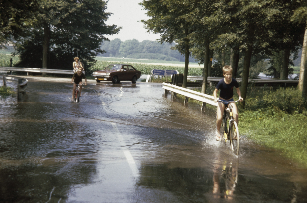 Eine Gruppe von Menschen, die auf Fahrrädern eine überschwemmte Straße entlangfahren, mit einem daneben fahrenden Auto, gesäumt von Geländern, Bäumen und Pflanzen unter einem klaren blauen Himmel.