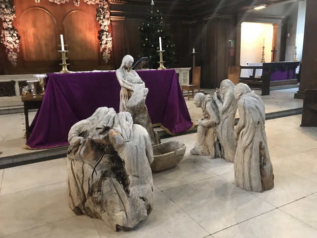 Krippe in einer Kirche mit Statuen auf dem Boden, einem Tisch mit einer Decke und Kerzenständern, Stühlen, einem beleuchteten Weihnachtsbaum und einer Wand im Hintergrund.