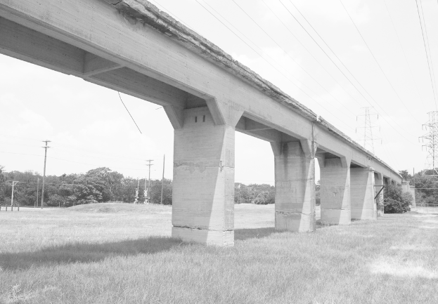 Eine Betonbr├╝cke auf einer gr├╝nen Wiese mit einem gro├čen elektrischen Mast und Kabeln ├╝ber ihr, umgeben von B├Ąumen und weiteren elektrischen Masten im Hintergrund.