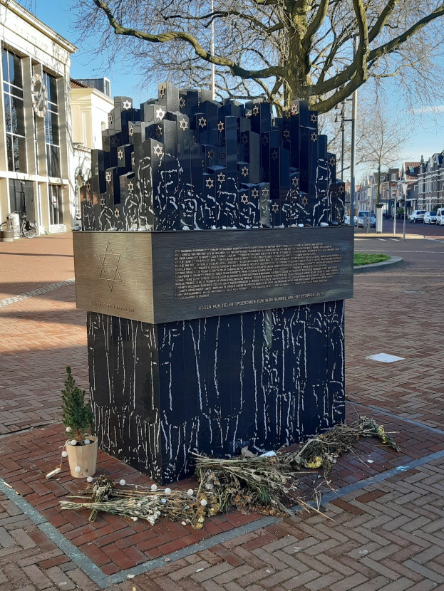Denkmal in einem Stadtplatz, das Holocaust-Opfern gewidmet ist, mit einem Baum davor, einem Topf auf dem Boden, verstreuten trockenen Blättern und im Hintergrund Gebäude, Fahrzeuge, Mäste und Bäume.