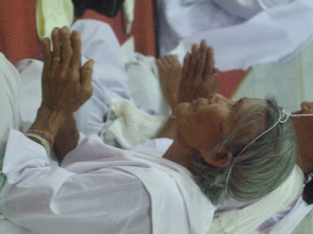 Eine ältere Frau liegt in einem Krankenhausbett mit gefalteten Händen, die besorgt mit geschlossenen Augen und auf den Händen ruhendem Kopf daliegt, umgeben von ein paar Menschen im Hintergrund.