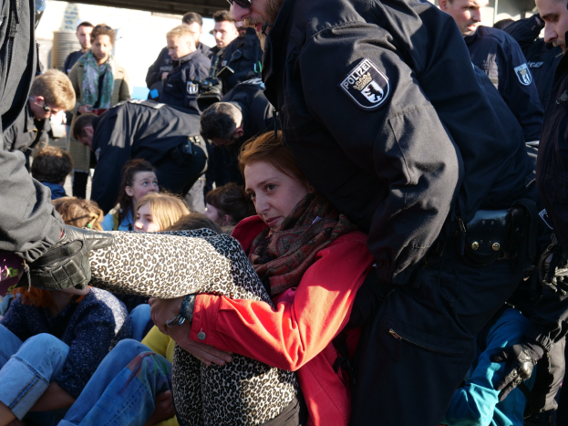 Eine Frau in dunkler Kleidung wird von einem Polizeibeamten in Uniform eskortiert, während eine Gruppe von Menschen hinter ihnen sitzt und steht, in der Nähe eines Schuppens unter einem klaren blauen Himmel.