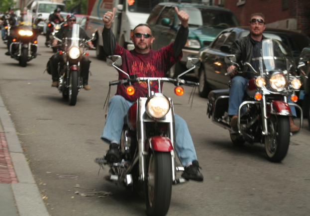 Eine Gruppe von Menschen auf Fahrrädern auf einer Straße, mit einer Person in einem roten Hemd, die ohne Hände fährt, einer Person in einer schwarzen Jacke, die lächelt, und zwei Autos und einem Van im Hintergrund.