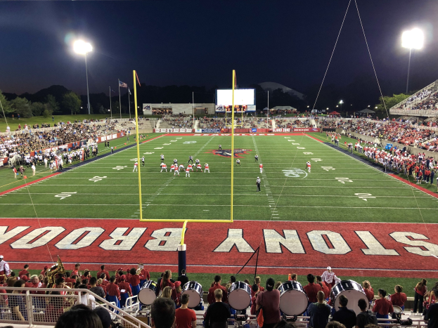 Ein nächtliches Fußballspiel in einem Stadion mit Spielern auf dem Feld, Zuschauern auf Stühlen und Musikern mit Instrumenten, umgeben von einem Metallzaun mit Laternen, Fahnen, einem Bildschirm, Bäumen, Hügeln und einem sternenklaren Himmel im Hintergrund.