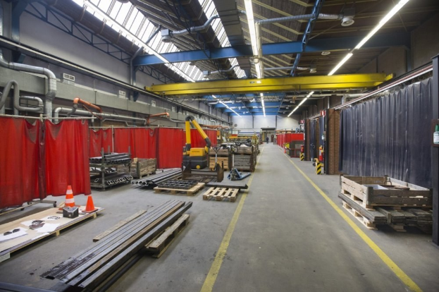 Großer Lagerraum mit Holzpaletten, Metallobjekten, Verkehrskegeln, Maschinenfahrzeug und roter Vorhang, Deckenrohre, Lampen und einer Vorhangwand auf der rechten Seite.