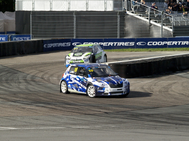Zwei Autos auf einer Rennstrecke mit Maschendrahtzaun und Bannern, umgeben von Zuschauern auf einer Treppe mit Geländer, mit Bäumen und Zelten im Hintergrund.