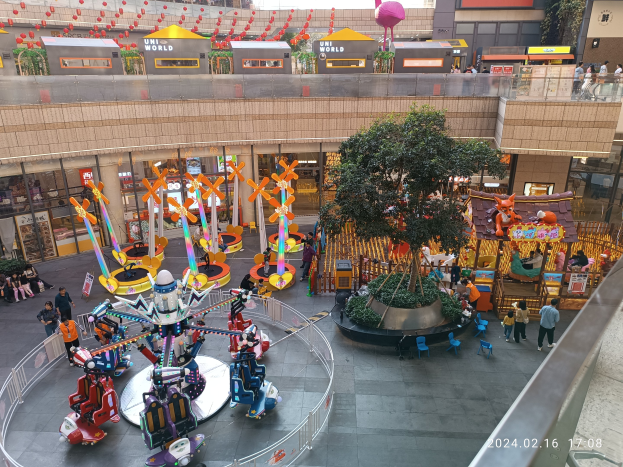 Ein Vergnügungspark in einem Einkaufszentrum mit verschiedenen Attraktionen, Bäumen, Menschen, Stühlen, Buden und Gebäuden sowie einem Wasserzeichen in der rechten unteren Ecke.