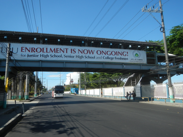 Ein Bus fährt unter einer Brücke mit einem Schild, auf dem "Jetzt Anmeldung" steht, während Menschen auf einem Fußweg darunter gehen, mit Strommasten und Drähten an der Straße und Bäumen und einem klaren blauen Himmel im Hintergrund.