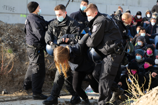 Gruppe maskierter Personen in Kapuzenpullis, einige bewaffnet, umringt eine Frau, die von der Polizei eskortiert wird, in einer städtischen Umgebung mit Pflanzen, Steinen und protestbezogenen Graffiti an einer Wand.