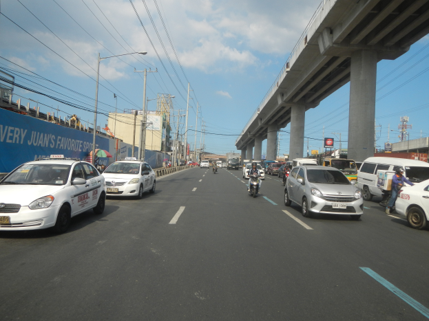 Eine belebte Hauptverkehrsstraße mit Autos und Motorrädern, eine Brücke im Hintergrund, Strommasten mit Drähten, Laternen, Gebäude und ein bewölkter Himmel.