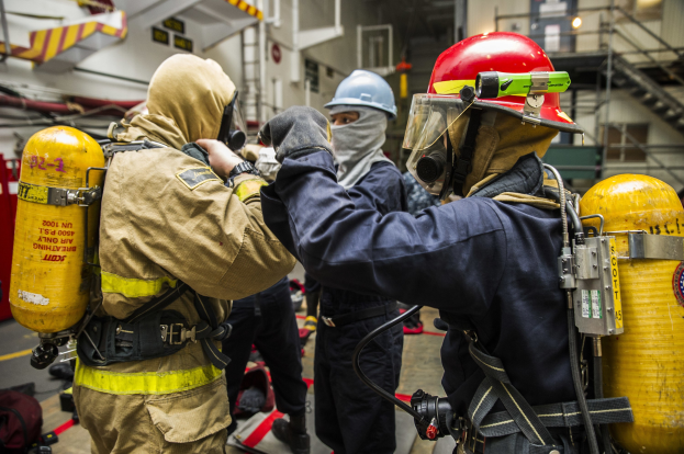 Feuerwehrleute in Schutzausrüstung mit Helmen, Handschuhen und Sauerstoffflaschen stehen zusammen, mit Taschen auf dem Boden und Equipment im Hintergrund.