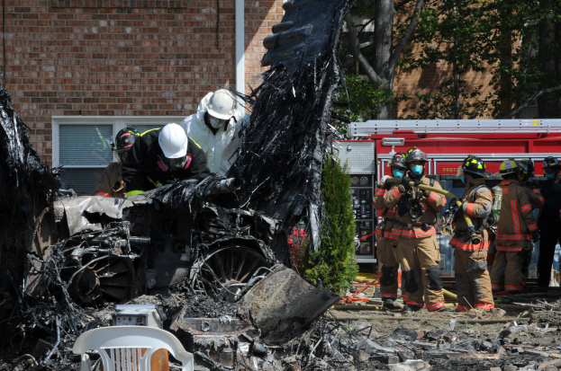 Feuerwehrleute in Schutzausröstung löschen ein Gebäude Feuer, mit verkohlten Trömmern, darunter ein Fahrzeug, Stuhl und Pflanzen, die auf dem Boden sichtbar sind.