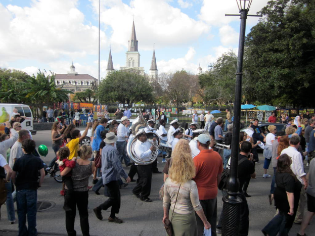 Eine Gruppe von Menschen auf einer Straße, einige spielen Musikinstrumente, umgeben von Gebäuden, Bäumen, Pflanzen, Zäunen, Fahrzeugen und Laternenmasten.