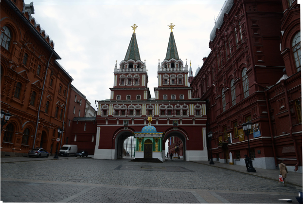 Fahrzeuge auf der linken Straßenseite, eine Person mit einer Tasche auf der rechten Straßenseite, Gebäude auf beiden Straßenseiten sowie Architektur und Wolken im Hintergrund.