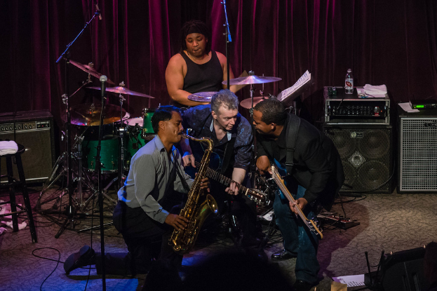 Eine Gruppe von Menschen auf der Bühne, die verschiedene Musikinstrumente spielen, mit einem Soundbox und einem Musiksystem dahinter.