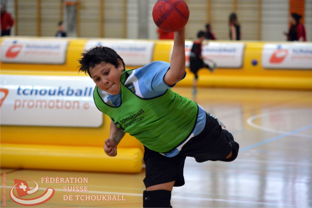 Ein Junge in einem weißen Hemd und dunklen Hosen wirft mit seiner linken Hand einen Ball, mit einem gelben Luftballon und mehreren Menschen im Hintergrund.