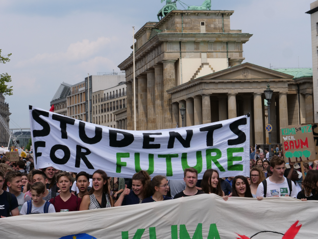Eine Gruppe von Schülern marschiert in Berlin, hält eine bunt bemalte Plakette, auf der "Students for Future" steht, mit Gebäuden, Bäumen und Himmel im Hintergrund.