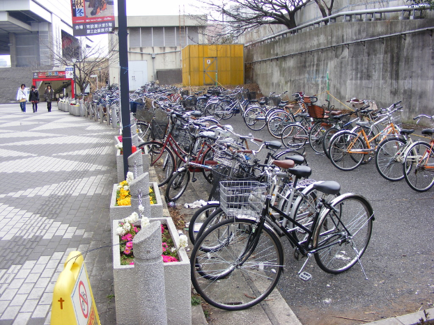 Im Vordergrund fahren Fahrräder auf der Straße nach rechts, während links drei Menschen auf einem Gehweg mit Geländer, Pfahl, Blumen und einem Schildtafel gehen. Der Hintergrund zeigt Gebäude, Bäume, eine Unterkunft, eine Wand und ein Banner oben.