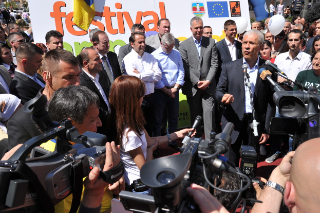 Eine Gruppe von Menschen, die auf einer Straße stehen und Kameras und Mikrofone halten, mit einem Plakat, Bäumen, Fahrzeugen, Topfpflanzen, einem Ballon, Pfosten und Gebäuden im Hintergrund.