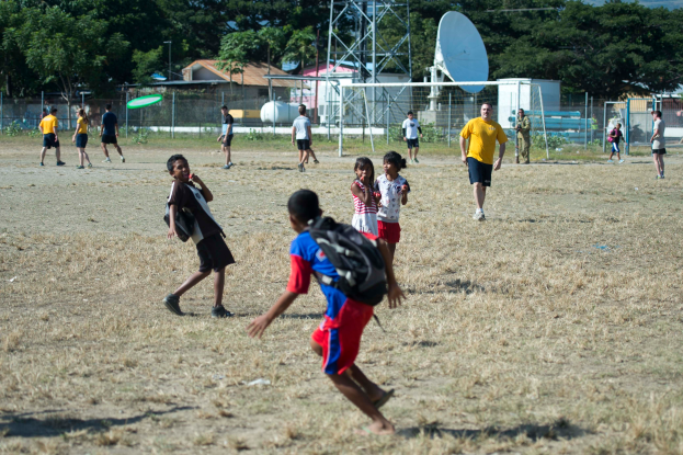 Eine Gruppe von Kindern, die auf einem von einem Metallzaun umgebenen Feld Fußball spielen, mit einer Schüsselantenne, Pfählen, einem Metallrahmen, Häusern mit Dächern, Pflanzen, Gras, Hügeln und einem bewölkten Himmel, wobei einige Kinder Taschen tragen.