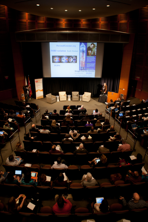 Ein mit Sitzreihen gefüllter Saal, in dem Menschen Gegenstände halten, beleuchtet von vielen Lichtern, mit Flaggen links, einem Podium rechts und einer Werbetafel links.