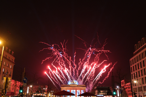 Eine belebte Straßenkreuzung in Berlin am Neujahrstag, voller Menschen, Autos und festlichen Dekorationen, mit Feuerwerk, das den Himmel und die Gebäude erhellt.