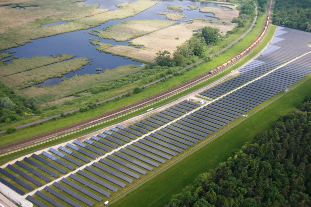 Luftaufnahme einer Solarpark mit Panelen, umgeben von Bäumen, Gras, Wasser und einer nahen Bahnlinie.
