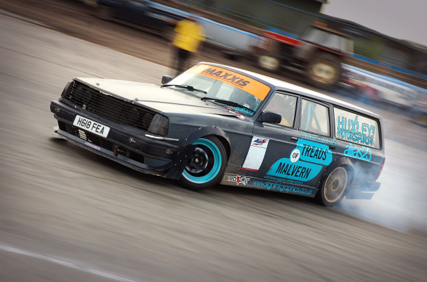 Ein Auto, das auf einer Rennstrecke mit Rauch ausfährt, umgeben von anderen Fahrzeugen und Zuschauern, mit einem unscharfen Hintergrund, der hohe Geschwindigkeit andeutet.