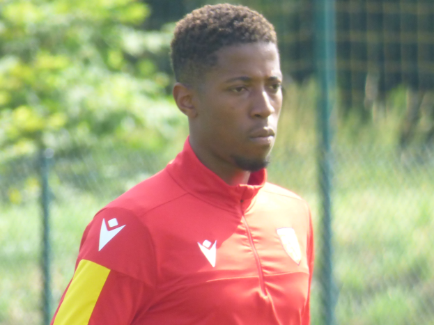 Ein junger Mann in einem roten und gelben Trikot steht auf einem Fußballfeld umgeben von einem Zaun und Bäumen und sieht entschlossen während eines Trainings.