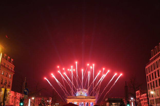 Eine belebte Straßen Szenerie in Berlin, Deutschland, mit dem Brandenburger Tor in der Mitte, beleuchtet von Gebäudelichtern und Feuerwerk am Nachthimmel.