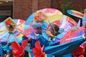 Eine Gruppe von Kindern in bunten Kostümen fährt auf einem Paradewagen, hält Schirme und hat ein Gebäude, Pflanzen und Lichter im Hintergrund.