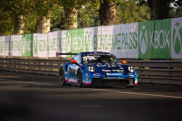 Blauer Porsche 911 GTS auf einer Rennstrecke mit einem Zaun und Bannern im Hintergrund, Bäume und einen klaren blauen Himmel sichtbar.