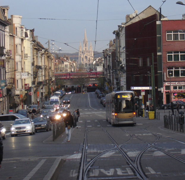 Eine belebte Stadtstraße mit Fahrzeugen, Fußgängern, einer Tram, Gebäuden, einer fernen Brücke, Bäumen und einem klaren blauen Himmel.