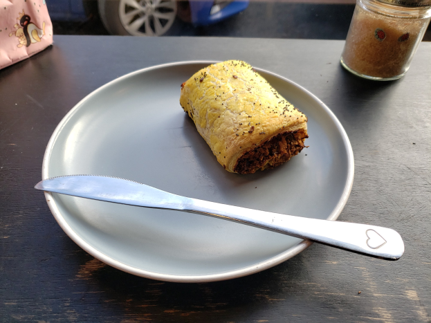 Ein Teller mit einem Stück Essen und einem Messer auf einem Tisch, ein Glasgefäß daneben und ein Fahrzeug durch die Scheibe im Hintergrund sichtbar.