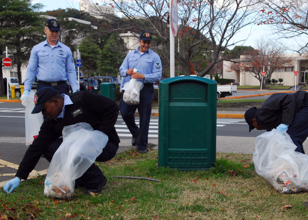 Drei Polizisten in Uniform sammeln Müll an einer Straßenseite, mit Bäumen, Gebäuden und Fahrzeugen im Hintergrund.