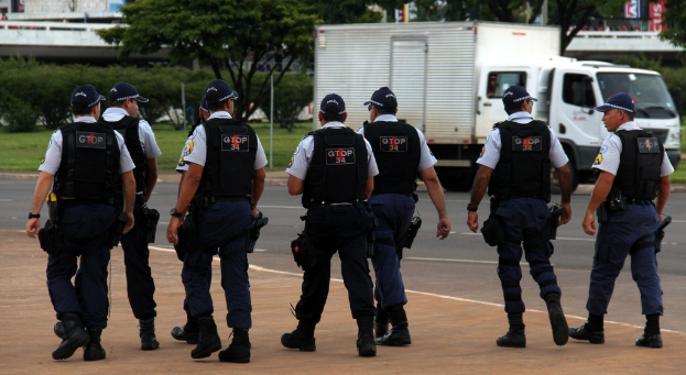 Eine Gruppe von Beamten in Uniform, einschließlich Mützen, Hemden, Schutzwesten, Uhren, Hosen und Schuhen, die auf einer Straße mit einem geparkten Lastwagen auf der rechten Seite neben einem Zaun gehen. Im Hintergrund sind Gebäude, Bäume, Pflanzen, Pfosten und Banner zu sehen, wobei in der oberen rechten Ecke eine Brücke zu erkennen ist.