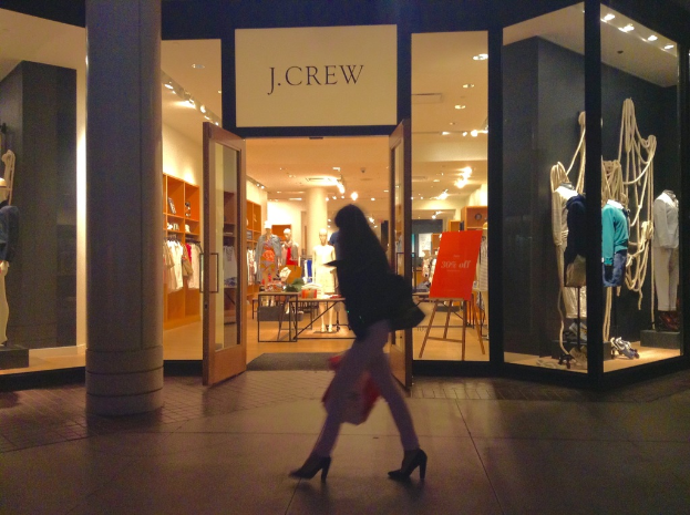 Ein Einkaufskomplex mit Glaswänden und -türen, mit Schaufensterpuppen, Tischen, hängenden Kleidern und Schränken, mit einer Frau, die daneben geht.