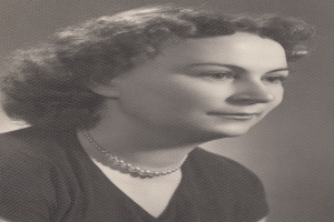 Ein Schwarz-Weiß-Foto einer Frau mit ernstem Gesichtsausdruck, deren Haar zurückgebunden ist und die eine Kette sowie ein einfaches Kleid mit hohem Kragen und langen Ärmeln trägt.