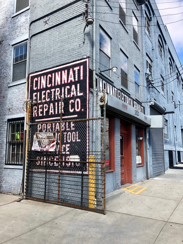 Das Gebäude der Cincinnati Electrical Repair Co. in Brooklyn, New York City, mit Fenstern, einer Tür, einem Schild, einem Metallzaun, einem Straßenpfosten und Drähten unter einem bewölkten Himmel.