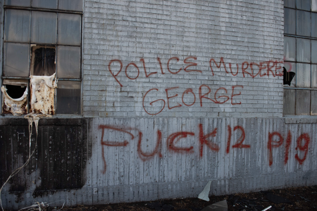 Ein Gebäude mit der Graffiti-Beschriftung "Polizei M"rdert George Fuck 12 P19" an der Seite, mit Fenstern und diversen Gegenst"anden vorne drauf.