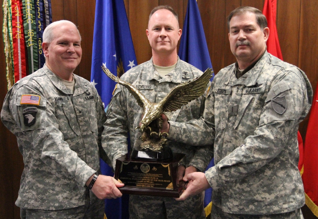 Drei Männer in Uniformen stehen mit Auszeichnungen, mit Fahnen dahinter und buntem Text in der oberen linken Ecke.
