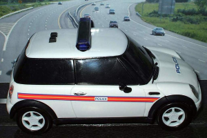 Ein weißer Polizeiwagen steht im Vordergrund, dahinter bewegen sich viele Autos auf der Straße, umgeben von Gras und Bäumen auf beiden Seiten.