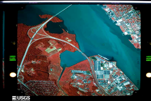 A satellite view of a city with a large body of water, illuminated by lights and surrounded by a dark background, with text at the bottom indicating it is a satellite image of the USGS.