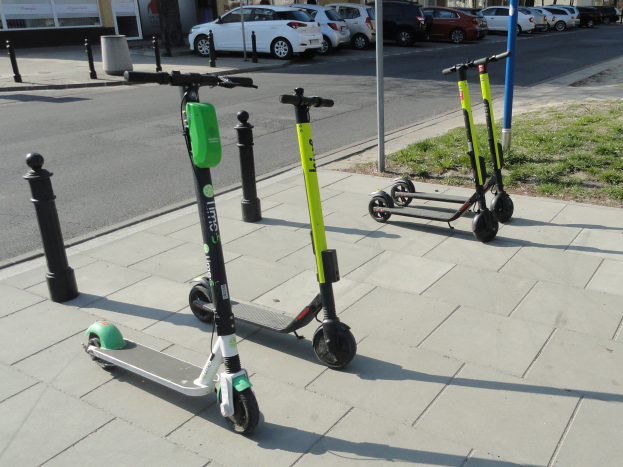 Drei Elektro-Scooter auf einem grasbewachsenen Straßenrand mit umgebender Infrastruktur und wolkenlosem Himmel.