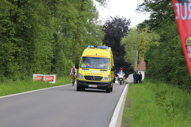 Ambulanz fährt auf Straße mit Fahrradfahrern daneben, gesäumt von Gras, Bäumen, Häusern, Strommasten und einem klaren blauen Himmel.